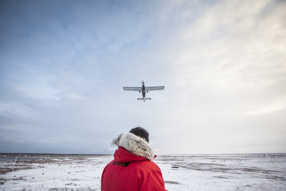 Person in red jacket outside during the day in Manitoba Hudson's Bay overlooking a small propeller plane takeoff